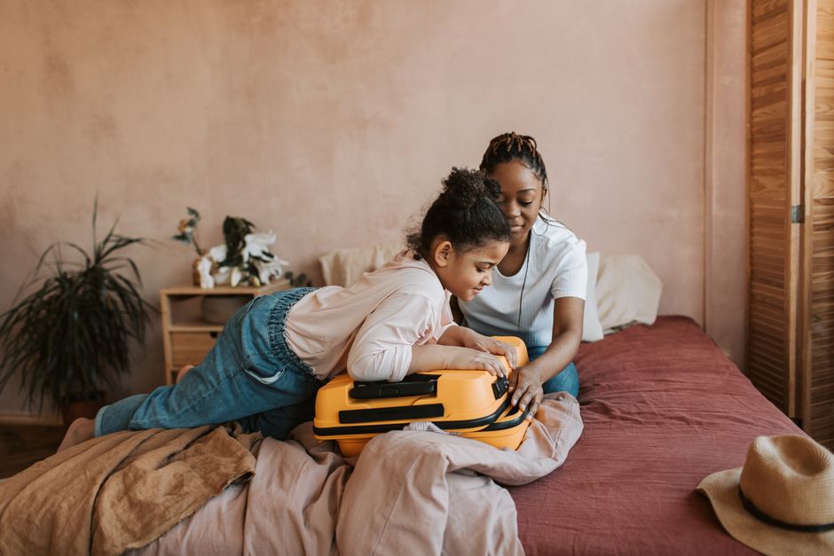 A mother and daughter pack a yellow suitcase in a cozy bedroom setting.