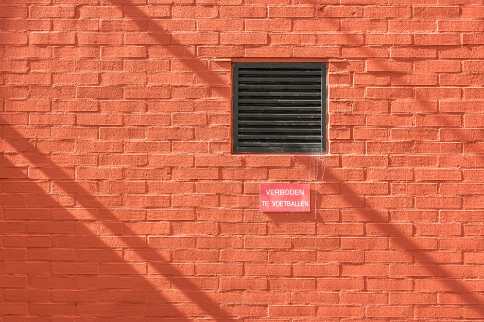 An orange brick wall featuring a black ventilation grate and a warning sign in Dutch.