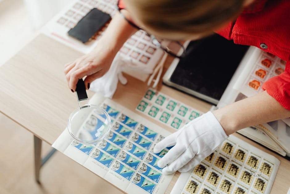 A person wearing white gloves inspects postage stamps using a magnifying glass.
