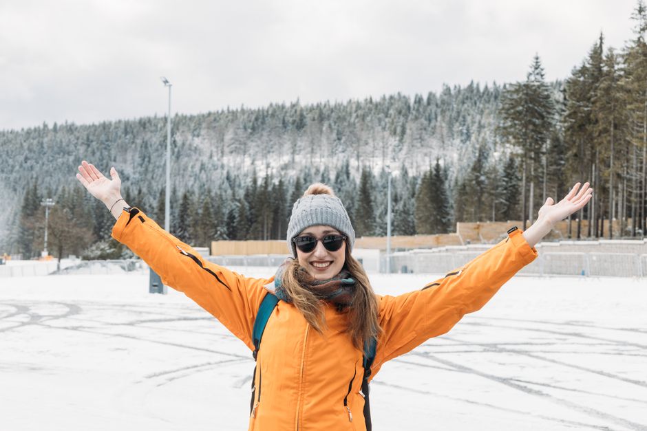 Smiling woman in orange jacket enjoying snowy outdoors, arms raised in winter forest.